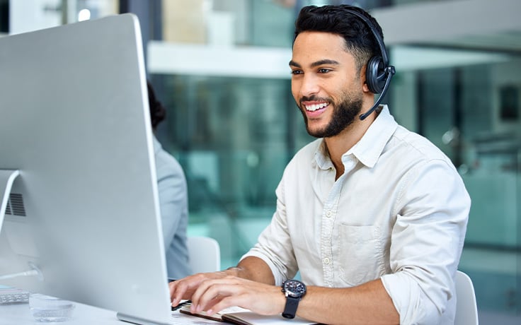A customer service representative with a headset is smiling and working on a computer in an office environment.