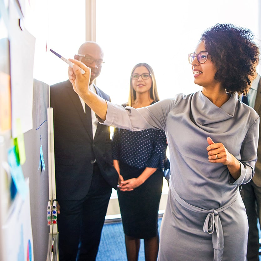 A business professional writing on a whiteboard while three other individuals in the background watch intently.