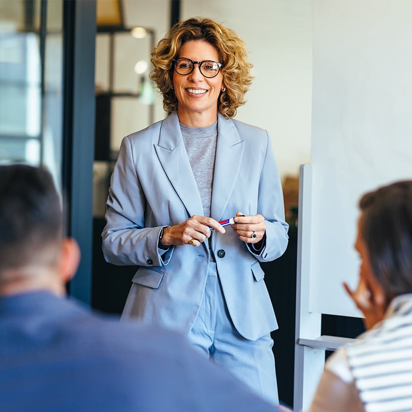A woman in professional attire is standing and speaking to a group of people who are seated and facing her in a business educational environment.