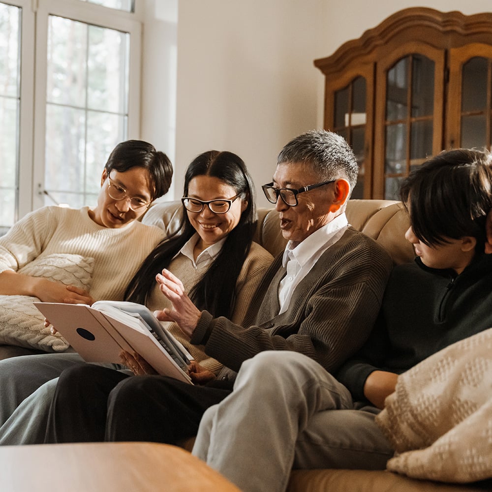 An older couple and two younger grandchildren sitting together on a sofa in a living room. The grandmother is holding open a photo album and the grandfather talking about the contents.
