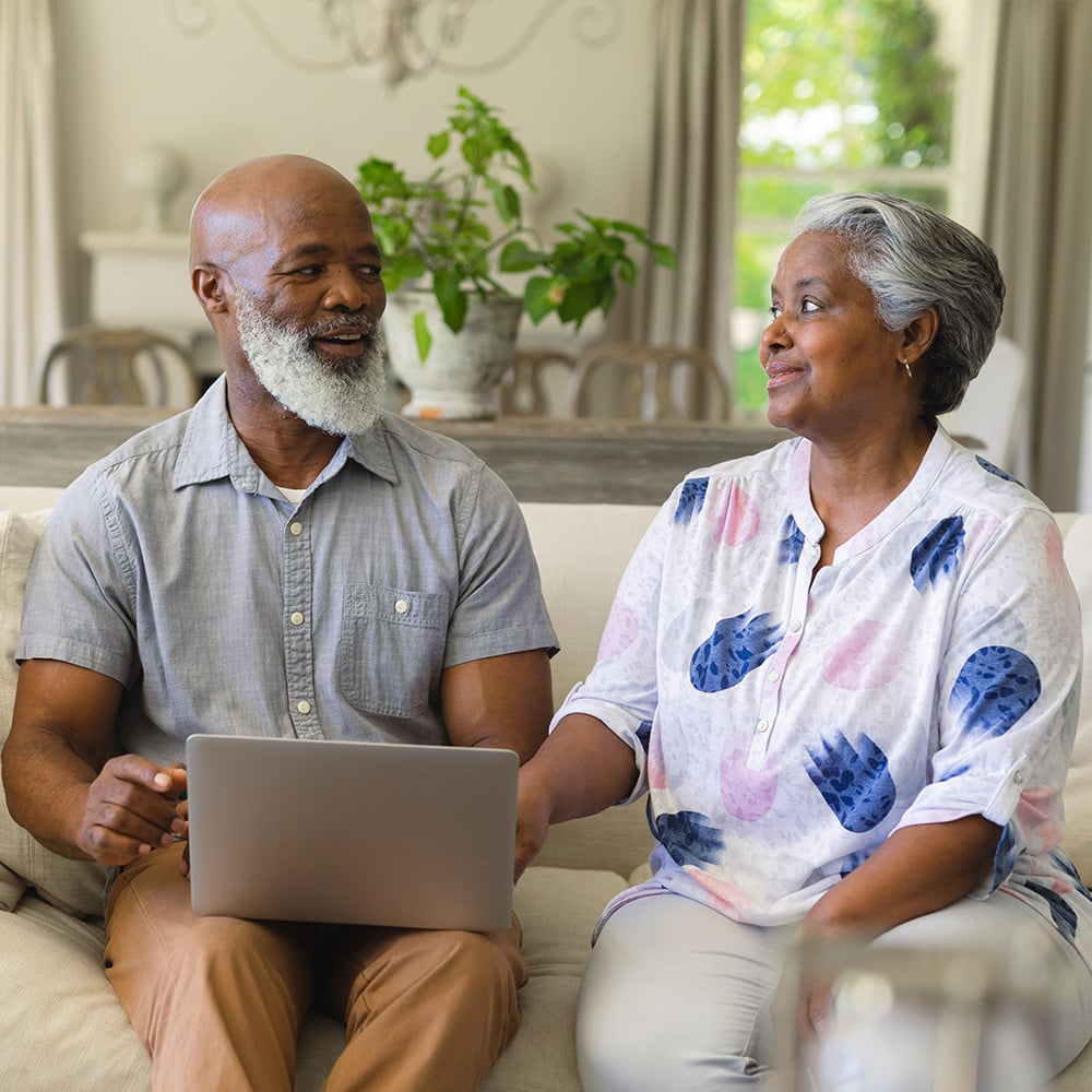 An older couple engaged in a pleasant discussion while sitting on a couch in home environment. A laptop is open in front of one person.