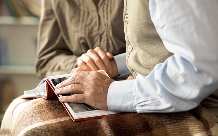A close-up of elderly and younger hands, clasped together atop a red photo album, suggesting an empathetic moment.