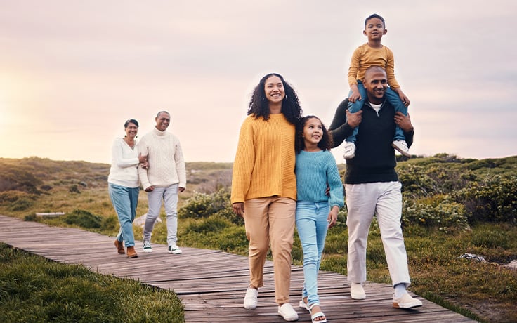 A multi-generational family of five is walking together on a boardwalk towards the sunset on a grassy terrain. The grandparents are farther behind the rest of the family, observing them from a distance.
