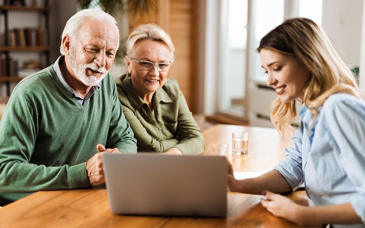 A funeral professional and an older couple are viewing ann open laptop together at a woden table, suggesting a collaborative or planning activity.