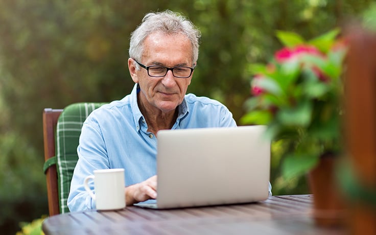 An older man sitting at a wooden table outdoors viewing a laptop open in front of him.
