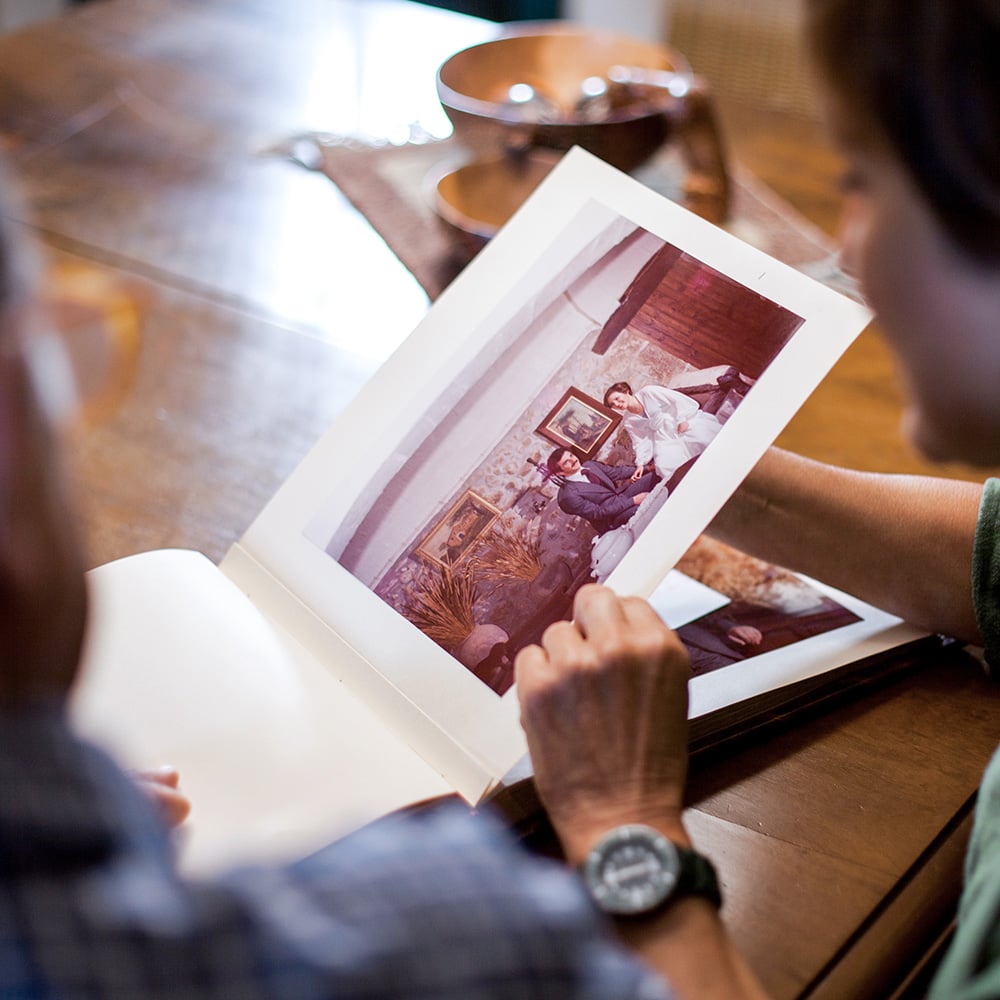 An older couple holding the photo album open to a page displaying a vintage photograph. The setting appears to be a cozy indoor environment, with a wooden table and a teapot or mug visible in the background.