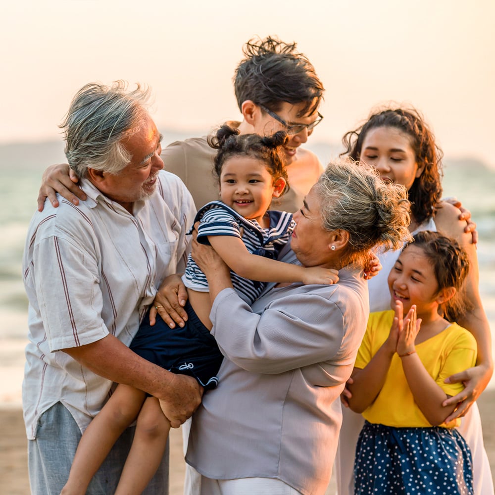 A multi-generational family, including grandchildren, adult children and grandparents, gathered close together, hugging and smiling on a beach.