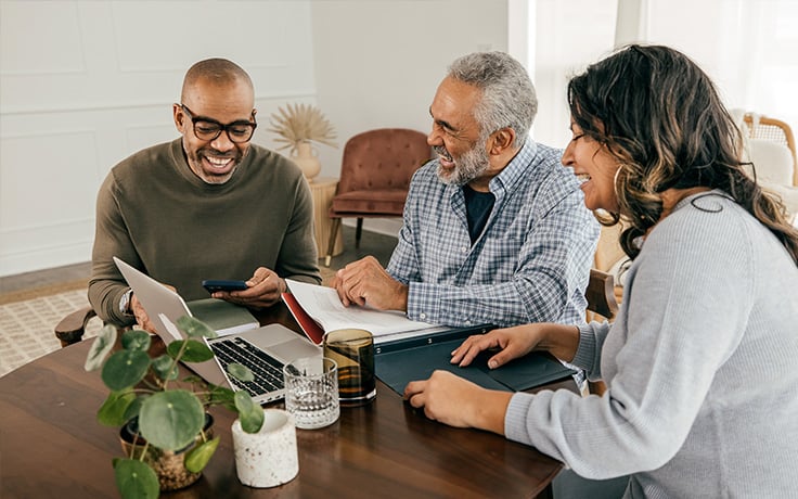 A friendly funeral professional and an older couple sitting at a wooden table with various office supplies, working together on a laptop.