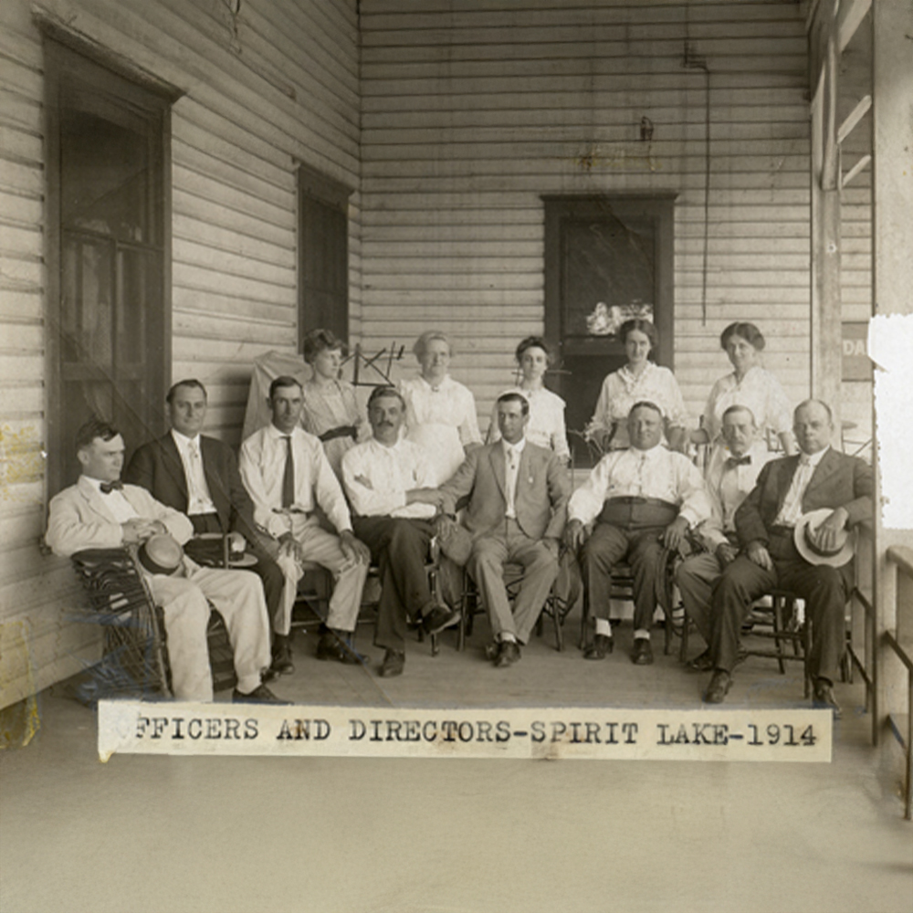 Historical black and white photograph of a group of men and women, officers and directors, posing for a group photo on a porch at Spirit Lake in 1914.
